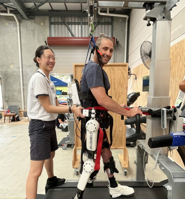 Man in exoskeleton device being supported by physiotherapy student.